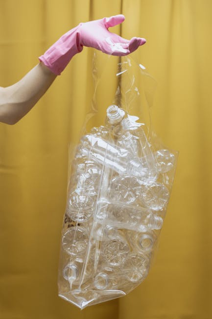 Close-up of several large black rubbish bags made of shiny plastic, tightly sealed and stacked against an off-white wall. The bags are placed on a paved surface, possibly in an outdoor setting such as