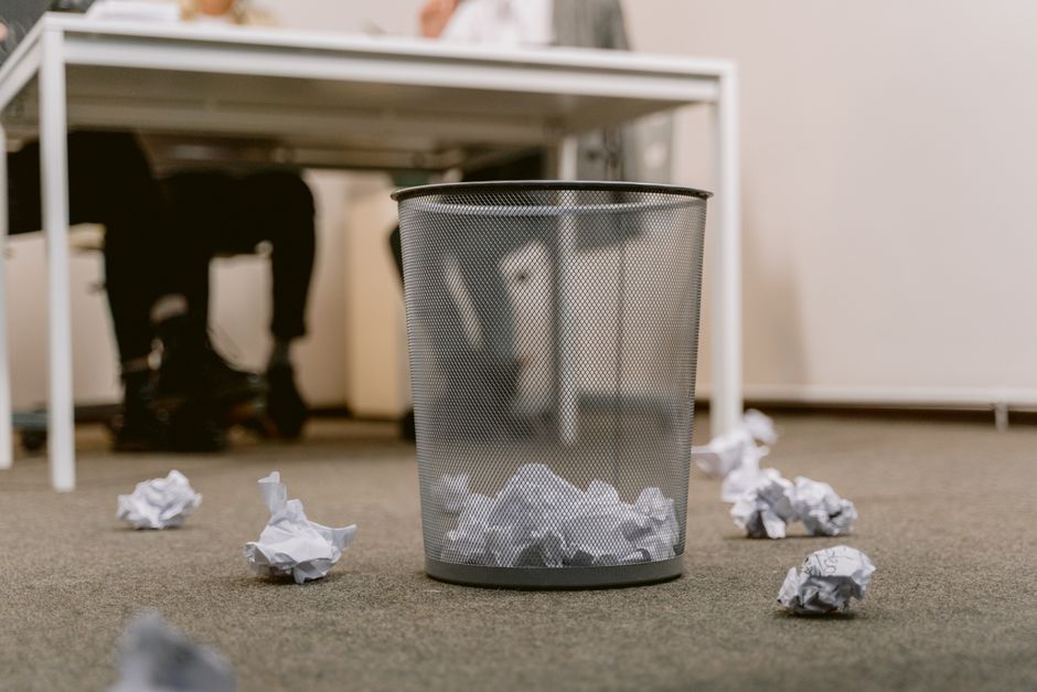 An office environment featuring a cylindrical metal mesh wastebasket placed on a gray carpeted floor, with crumpled white paper scattered around its base. In the background, a white desk with fours le