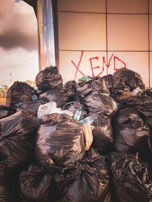 A waste collection operative wearing a red and yellow high-visibility jacket is standing beside a large, red rubbish truck parked on the side of a busy street. The rear of the truck, with its open com