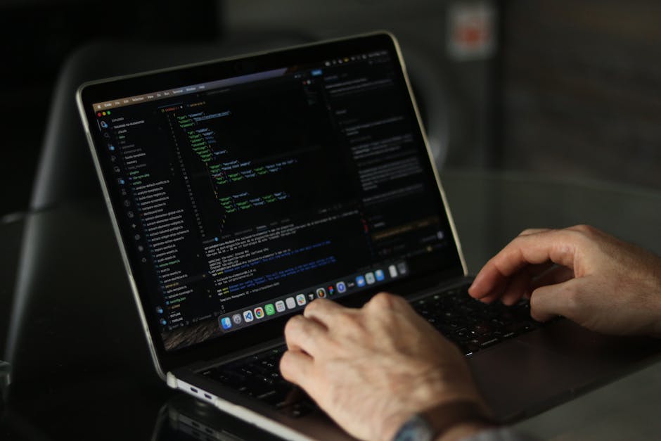 A man with short brown hair, glasses, and a beard is sitting at a wooden desk working on a computer with two large monitors. The monitors display lines of computer code with highlighted syntax on a da